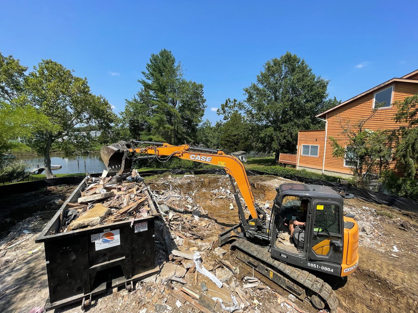 Excavator loading demolition debris into a dumpster beside a waterfront home